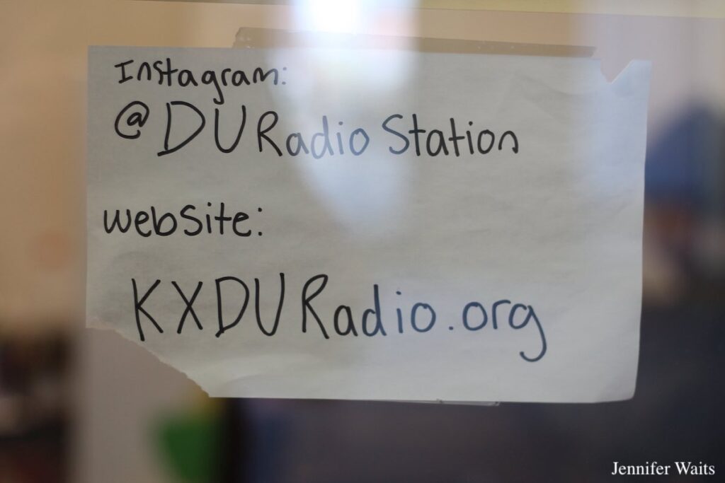 Photo of sign at college radio station KXDU. Sign reads: Instagram: @DUradioStation website: KXDURadio.org