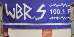 Photo of banner for college radio station WBRS. Blue Banner reads WBRS 100.1 FM and is hanging on a wall. Photo: J. Waits